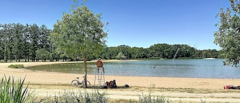 Un paysage au bord d’un lac, avec un vélo, une échelle et une personne assise sur le sable.