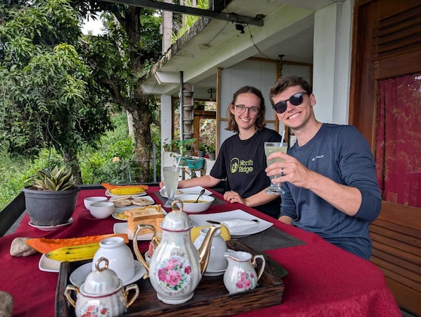 Two people seated at an outdoor table with a red tablecloth, various dishes, and a potted plant. - Dream Nest Ella (Ella)