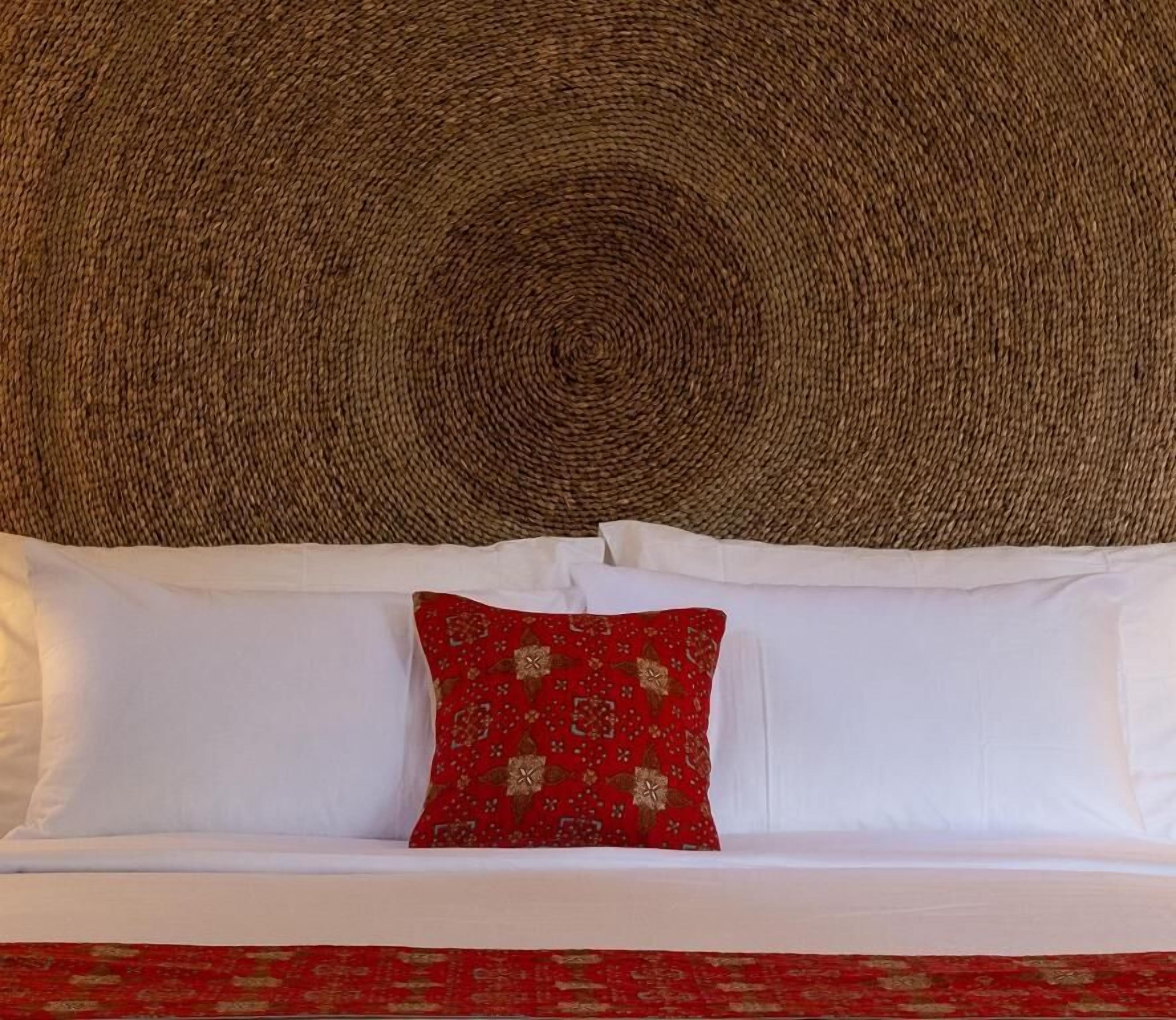 Una cama con ropa de cama blanca y roja, un cabecero tejido y una almohada estampada.
