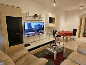Ein modernes Wohnzimmer mit einem wandmontierten Flachbildfernseher, einem Glastisch, beigen Sofas und einem Essbereich mit roter Tischdecke.