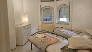 A bedroom with two beds, a bedside table, and a lamp. There are arched windows with blinds. - Nice small apartment for 2 in Kirkel with parking & quiet location (Kirkel)