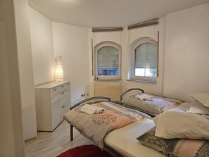 Ein Schlafzimmer mit zwei Betten, einem Nachttisch und einer Lampe. Es gibt gewölbte Fenster mit Jalousien.