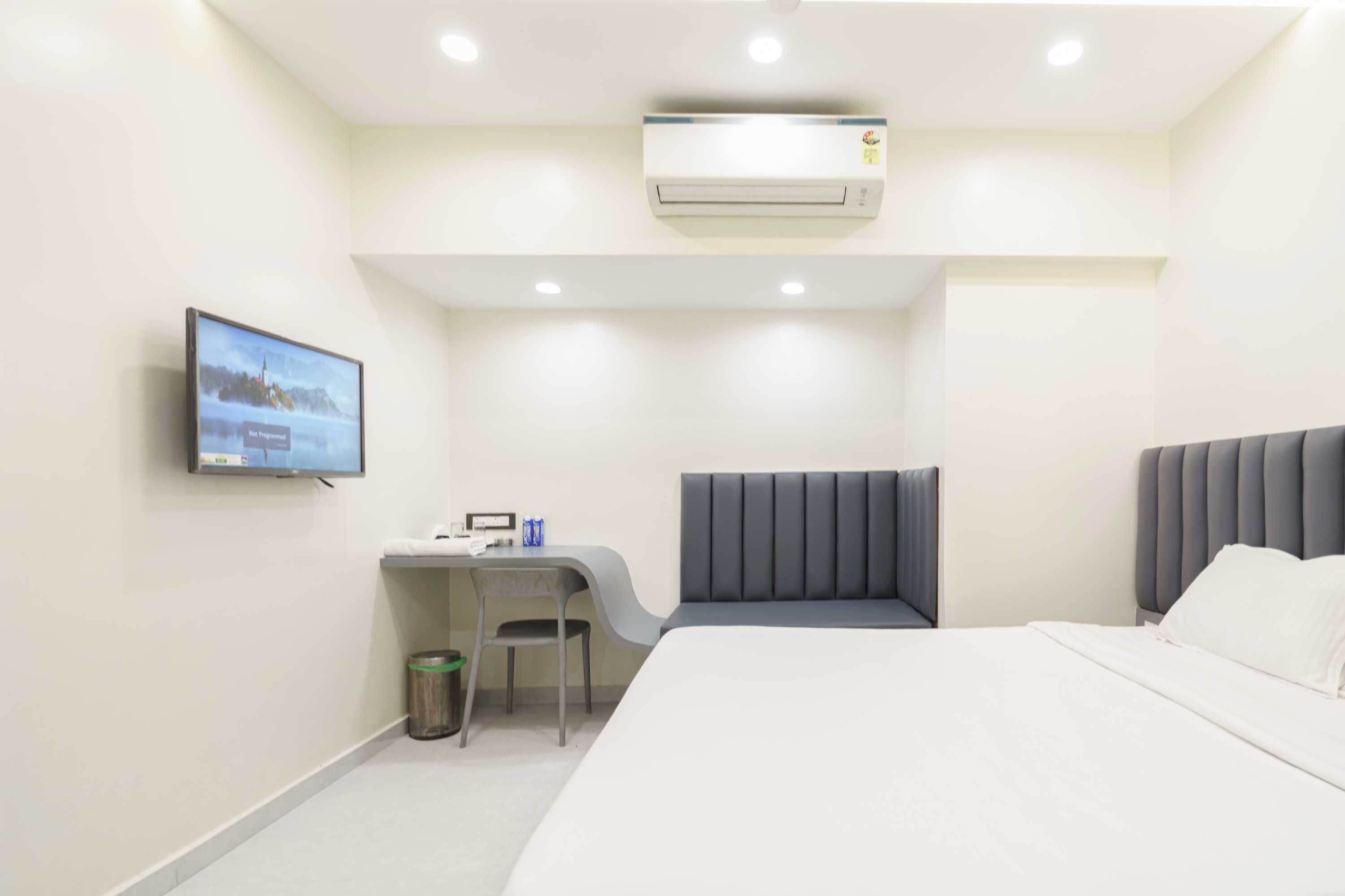 A hotel room with a bed, a desk with a chair, a television, and an air conditioning unit.