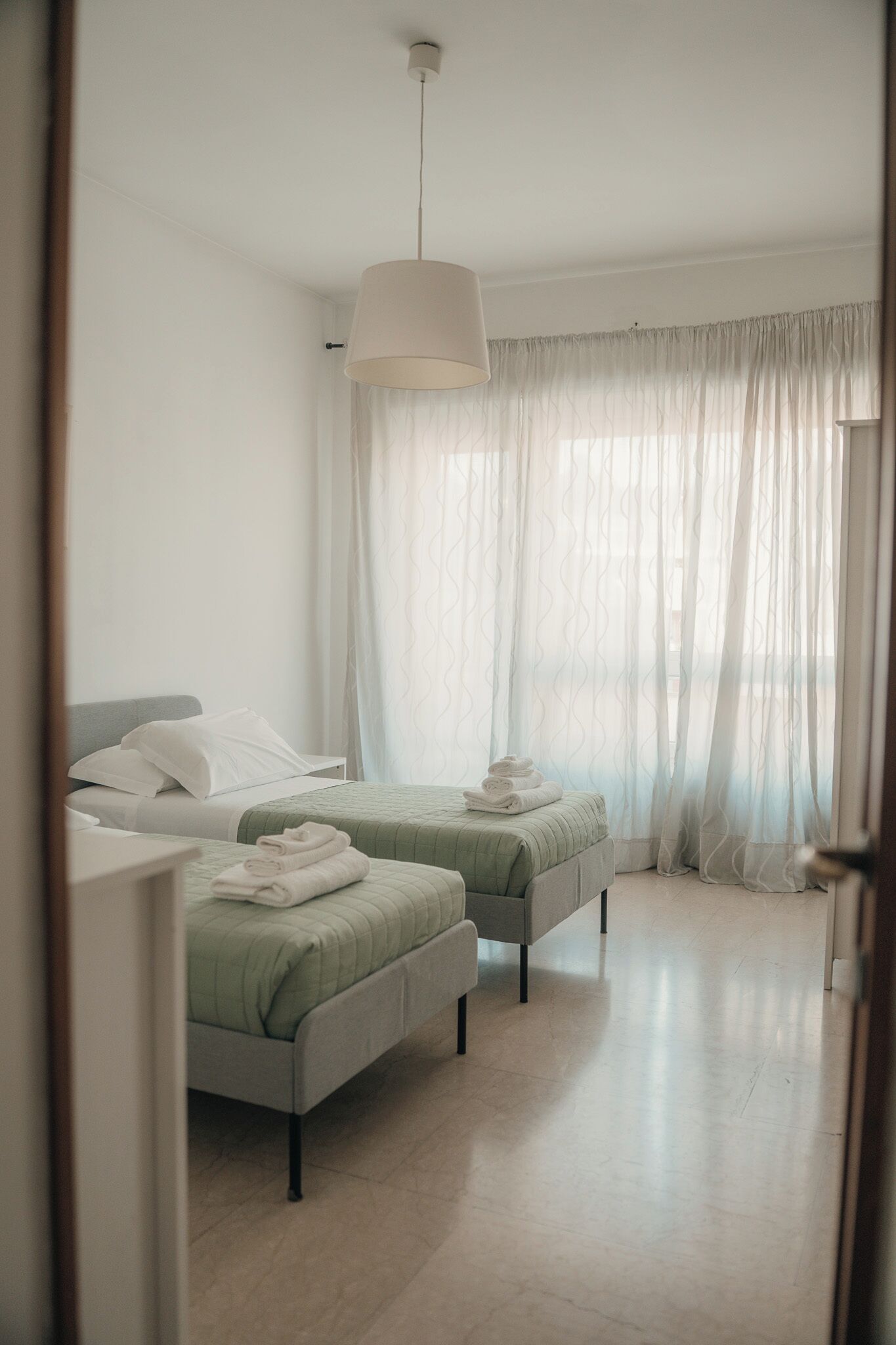 A hotel room with two beds, a hanging light fixture, sheer curtains, and a tiled floor.