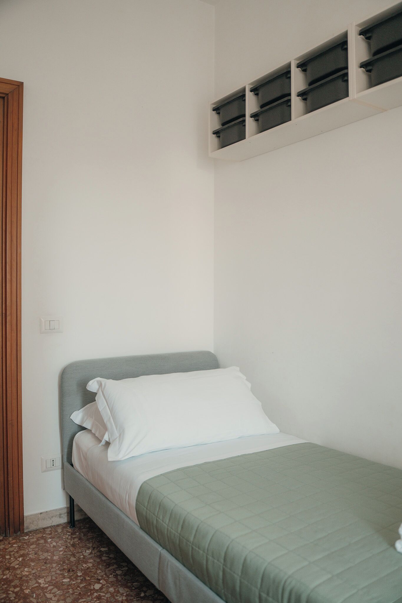 Ein Einzelbett mit grüner Bettdecke und weißen Kissen in einem Zimmer mit weißer Wand und Holztür.