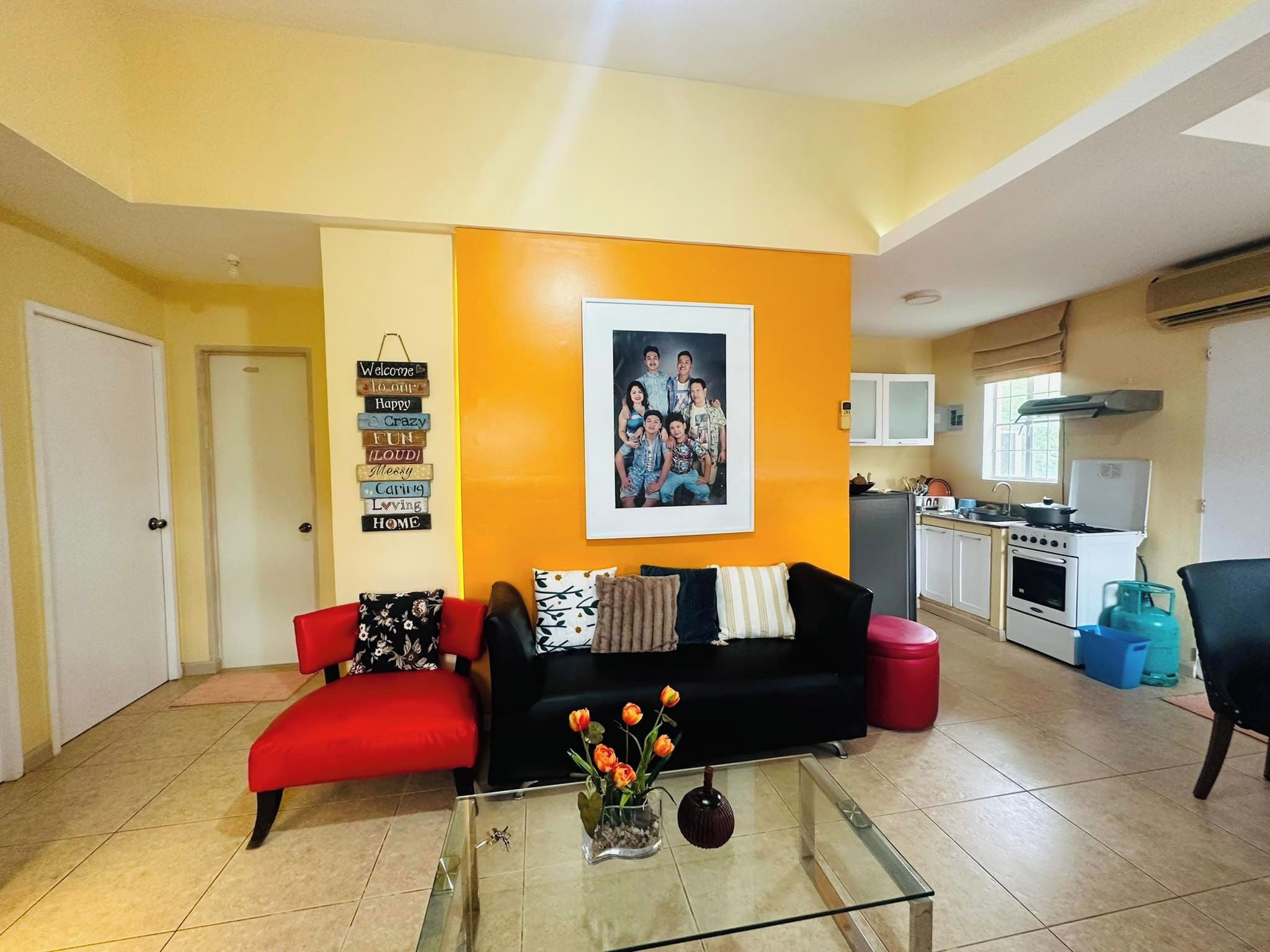 A living room with a black sofa, red armchair, and a glass coffee table with flowers. A framed photo is on the wall, and a sign with words like 'Welcome' and 'Home' is visible.