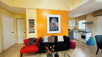 A living room with a black sofa, red armchair, and a glass coffee table with flowers. A framed photo is on the wall, and a sign with words like 'Welcome' and 'Home' is visible.