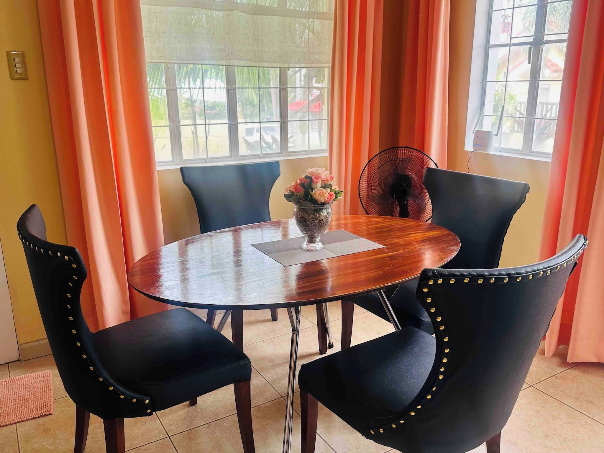 A round dining table with a vase of flowers, surrounded by black chairs with gold studs, in a room with orange curtains and a window.