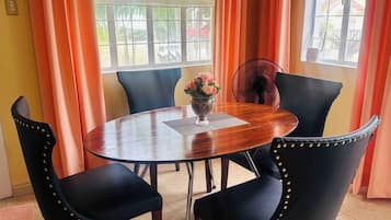 A round dining table with a vase of flowers, surrounded by black chairs with gold studs, in a room with orange curtains and a window.