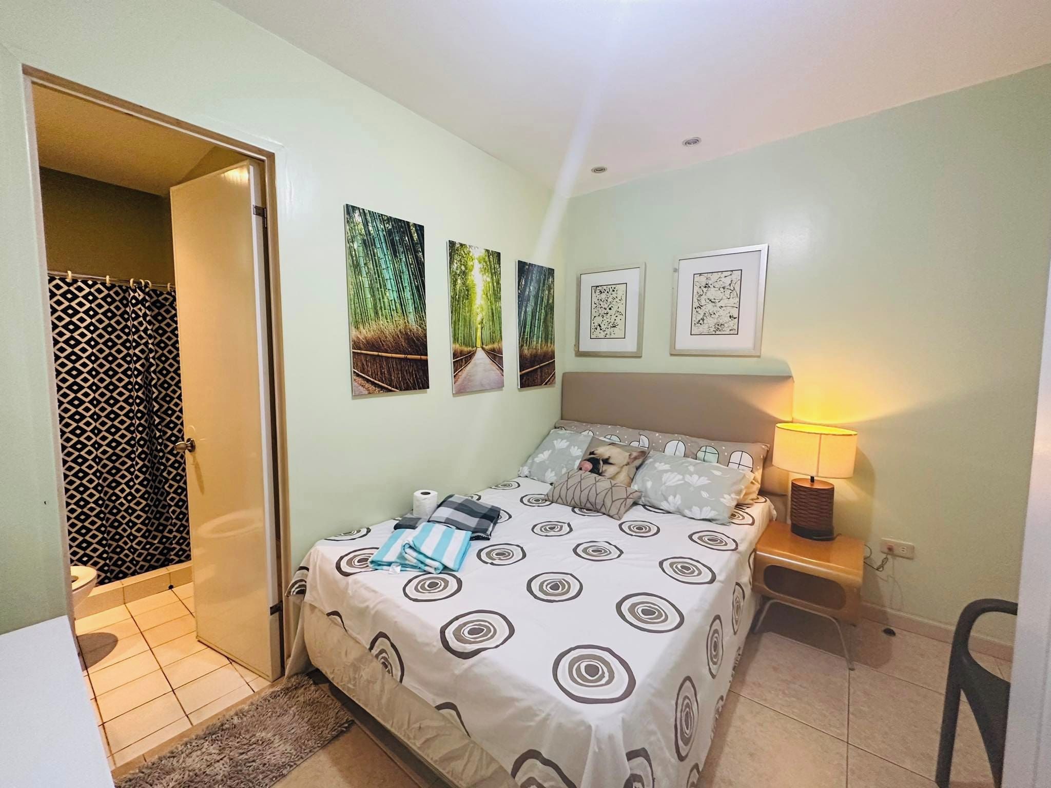 A bedroom with a bed, a bedside table with a lamp, a wall with framed pictures, and a bathroom with a shower curtain.
