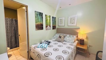 A bedroom with a bed, a bedside table with a lamp, a wall with framed pictures, and a bathroom with a shower curtain.
