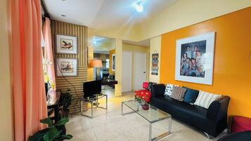 A living room with a sofa, coffee table, and a TV. There is a kitchen in the background.