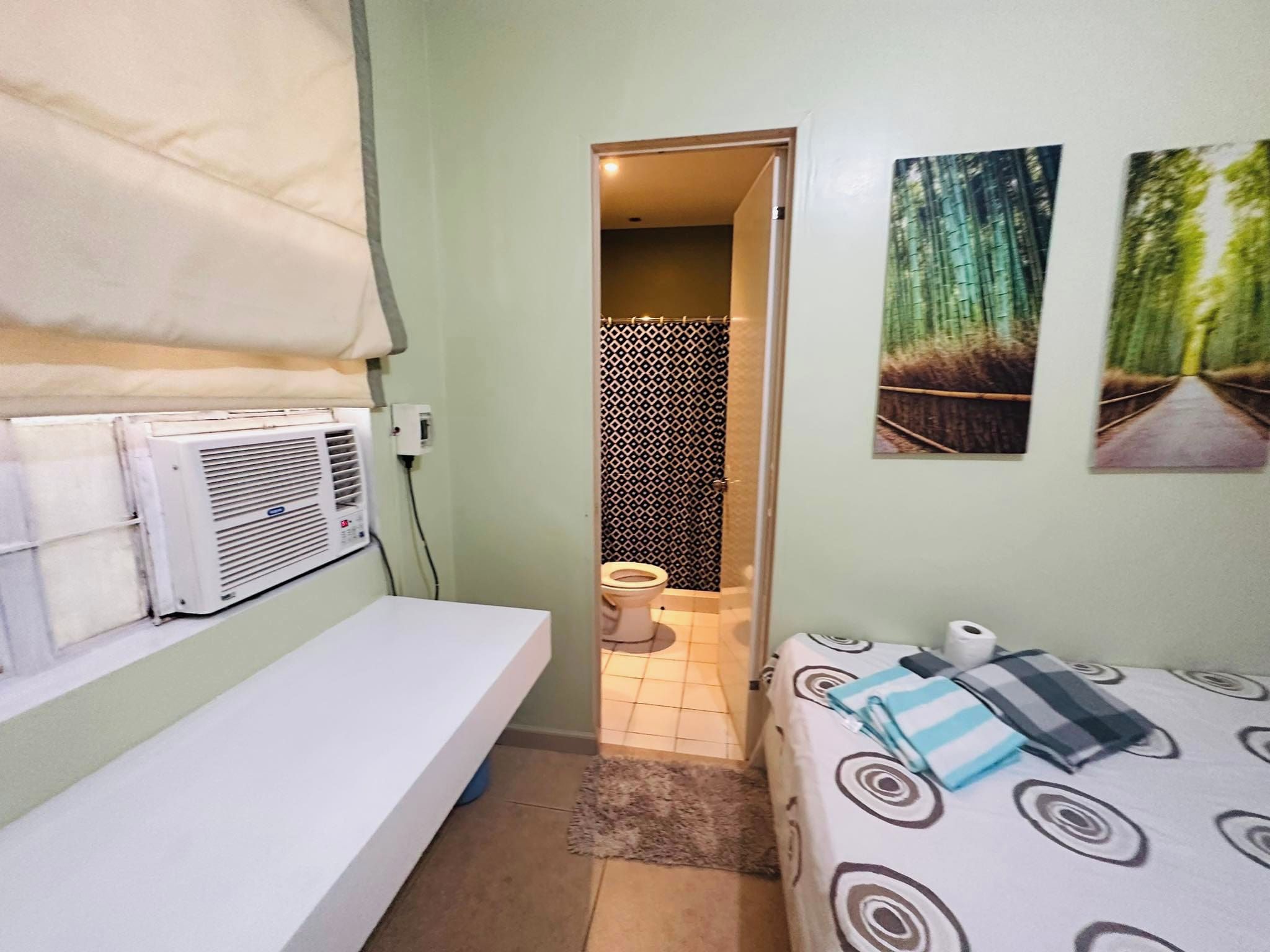 Una habitación pequeña y funcional con cama, mesita de noche, ventana con cortina, aire acondicionado instalado en la pared y un inodoro.