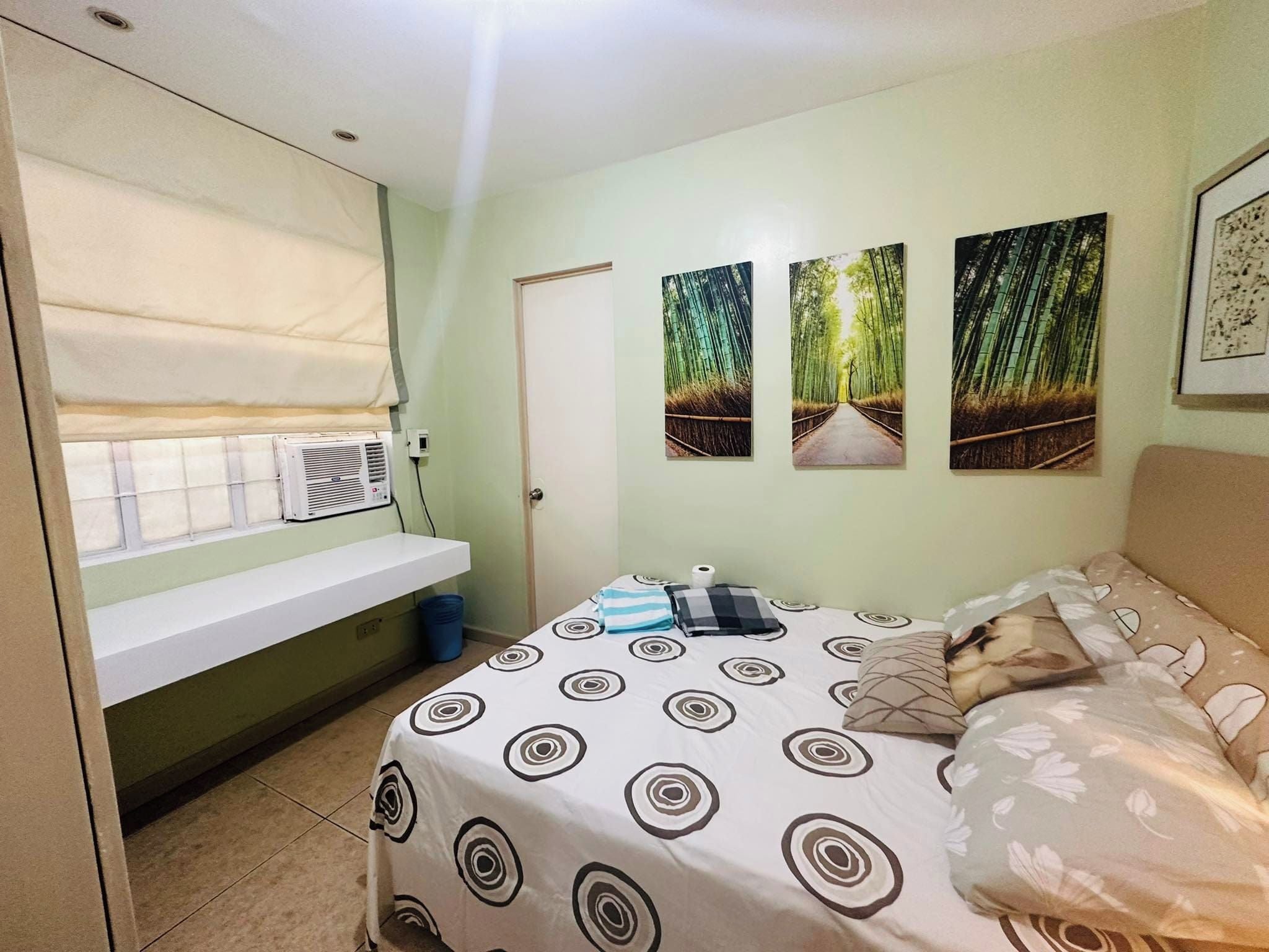 A bedroom with a bed, a window with blinds, a wall-mounted air conditioner, and three framed pictures.