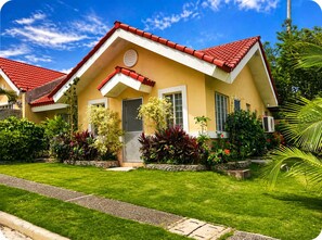 A two-story house with a red-tiled roof, a front porch, and a well-maintained garden with various plants and a lawn. - Charming 3-bedroom House Rental in Guarded Cagayan de Oro Suburb (Cagayan de Oro)