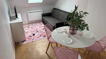 A small, modern apartment with a dining area featuring a round table and pink chairs, a grey sofa, a potted plant, and a radiator.