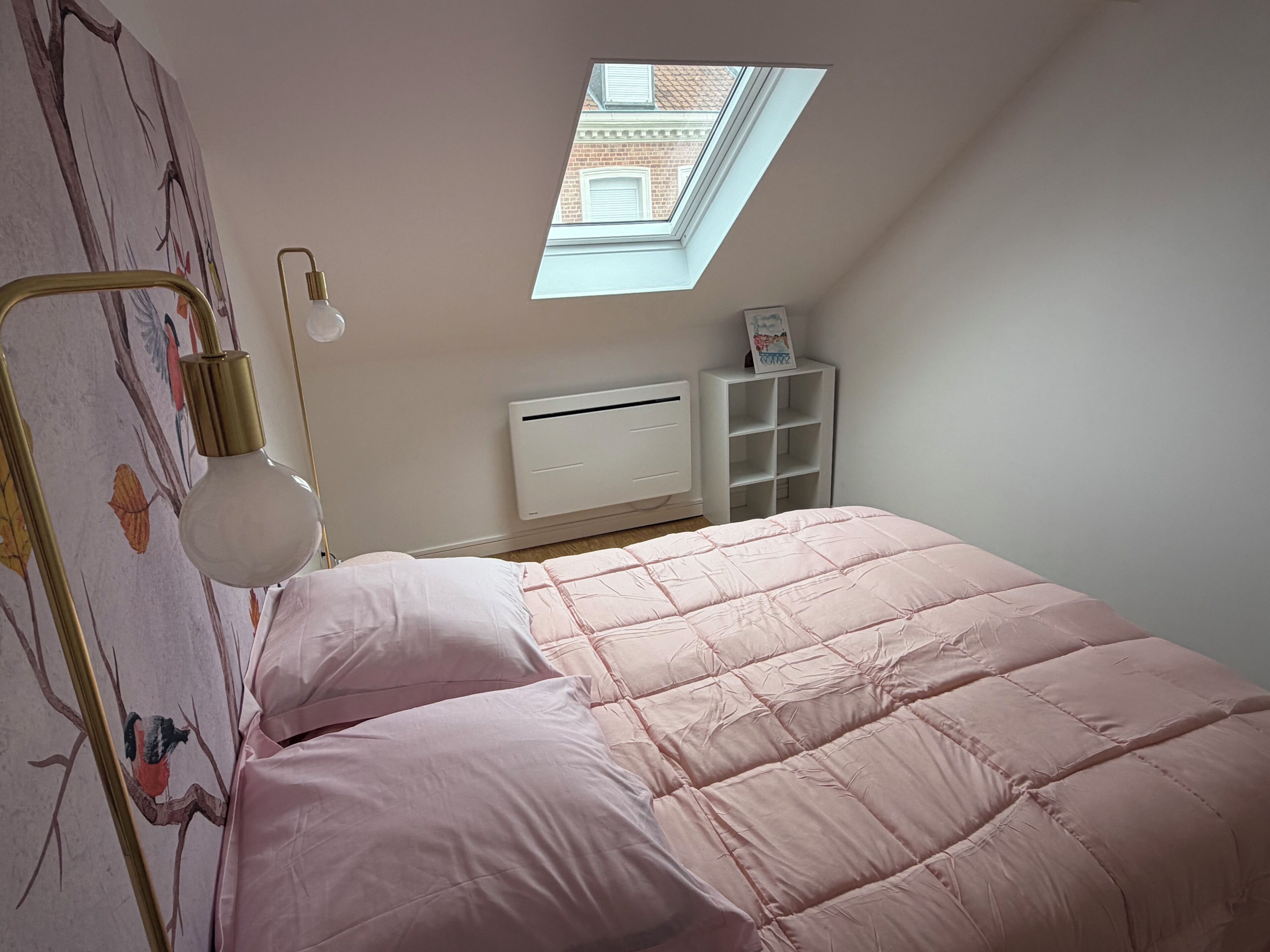 A bedroom with a sloped ceiling, a skylight, a bed with pink bedding, a bedside lamp, a radiator, and a white shelving unit.