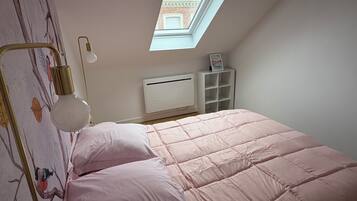 A bedroom with a sloped ceiling, a skylight, a bed with pink bedding, a bedside lamp, a radiator, and a white shelving unit.