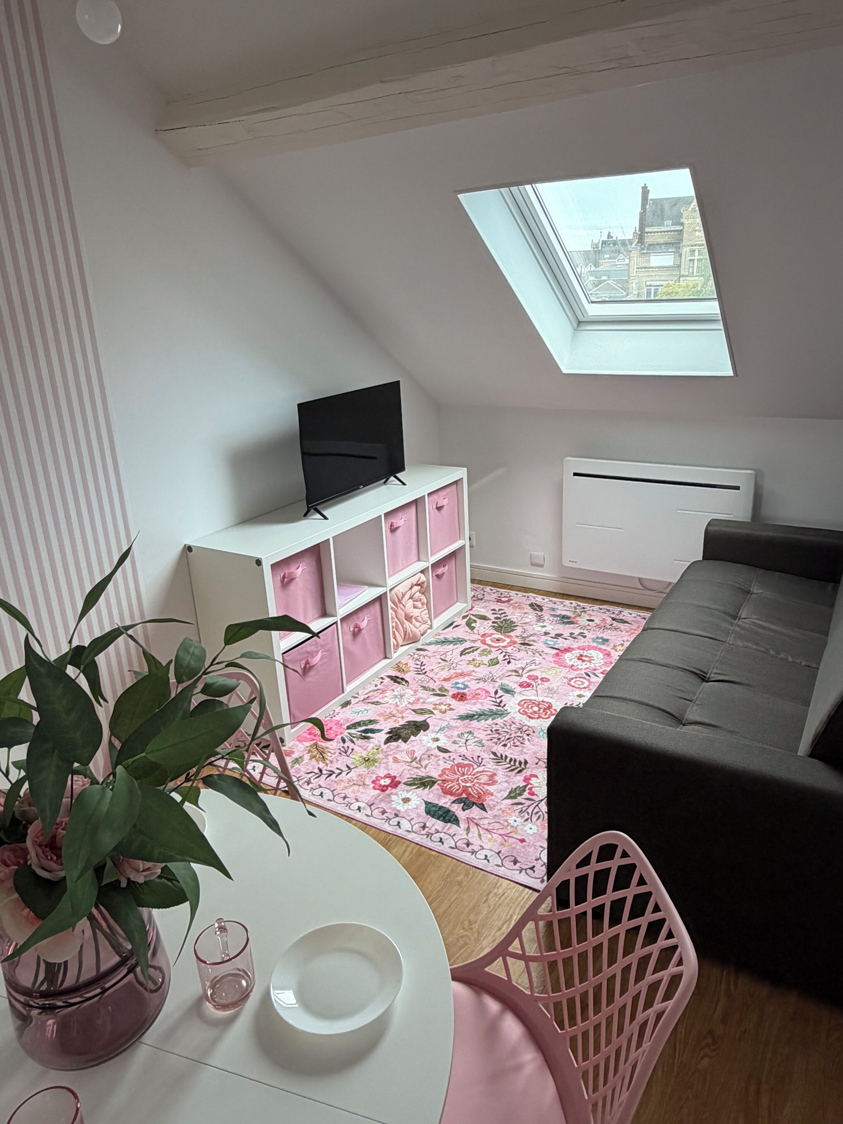 Una habitación con techo inclinado, un televisor sobre un mueble blanco, un tapete floral, un sofá gris y un sillón rosa, además de un tragaluz.