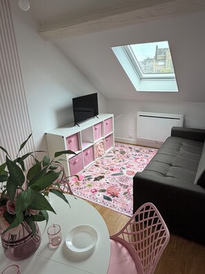 Una habitació amb sostre inclinat, una televisió sobre un moble blanc, una catifa floral, un sofà gris i una cadira rosa, i un finestral al sostre.
