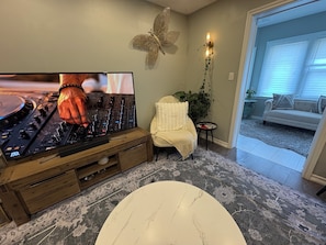 A living room with a DJ setup, a white armchair, a decorative wall piece, and a view into the adjacent room.