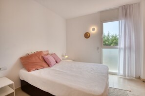 A bedroom with a bed, pillows, a bedside table, a lamp, and a view of trees through a window. - JULIA: Modern Apartment with Sea Views Next to Riells Beach (L'Escala)