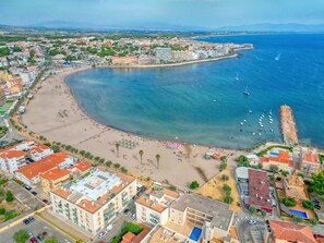 An aerial view of a coastal city with a curved bay, buildings, and a beach. - JULIA: Modern Apartment with Sea Views Next to Riells Beach (L'Escala)