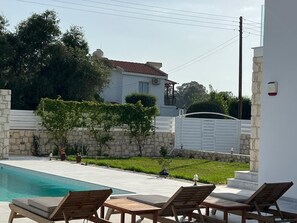 A pool area with lounge chairs, a stone wall, and a house in the background. - Jomavi Beach Eco-Villa (Polis Chrysochous)