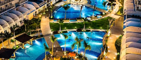 A multi-level hotel with a large central pool area, multiple smaller pools, and a well-lit outdoor lounge area with umbrellas.