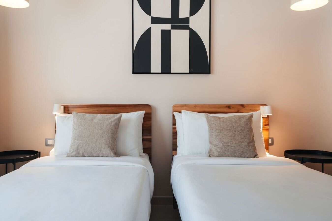 Et hotelværelse med to senge, hver med en træsenggegærd, hvidt sengetøj og en enkelt pude. Der er en billedkunst ophængt på væggen over sengene.