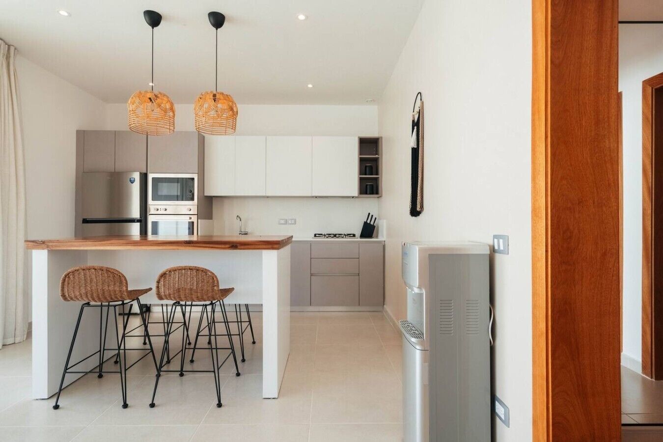 A modern kitchen with a central island, two wicker chairs, a refrigerator, and pendant lighting.