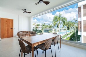 A modern dining area with a large table and wicker chairs, a wooden door, and a view of a pool and palm trees. - 2-Bed Ocean View on Beach • Sol de Arena C5 (Las Terrenas)