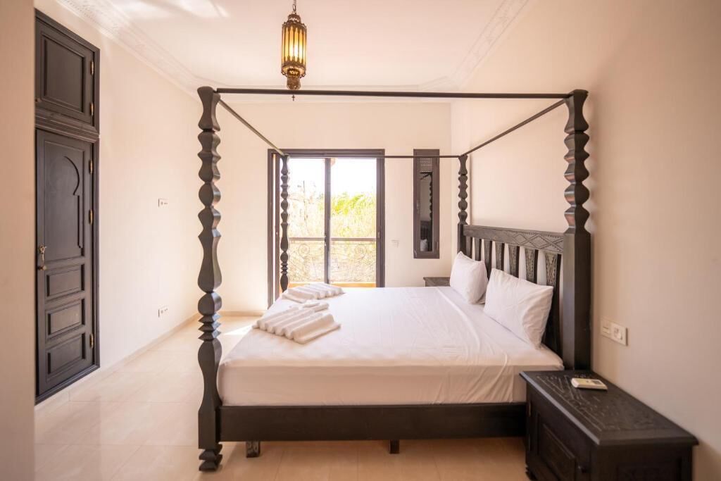 A four-poster bed with a dark wooden frame and white bedding, a bedside table with a lamp, and a view of the outdoors through a large window.