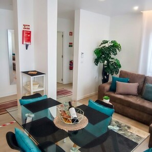 Ein modernes Wohnzimmer mit einem Glastisch, einem braunen Sofa mit Kissen, einer Topfpflanze und einem kleinen Tisch mit Flasche und Gläsern.