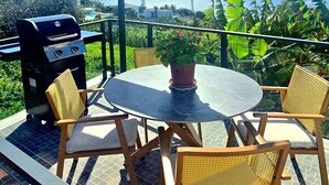 Une terrasse extérieure avec une table ronde, quatre chaises en osier, un barbecue et une plante en pot.