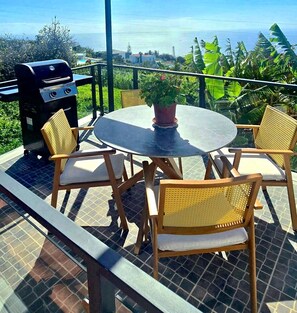 Eine Terrasse im Freien mit einem runden Tisch, vier Korbstühlen, einem Grill und einem Blumentopf.