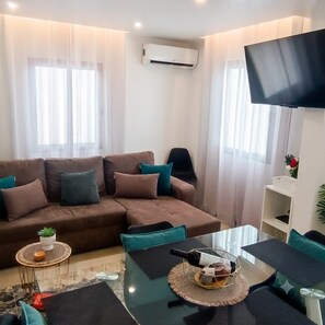 Ein Wohnzimmer mit einem braunen Sofa, türkisfarbenen Kissen, einem Glastisch und einem an der Wand befestigten Flachbildfernseher.