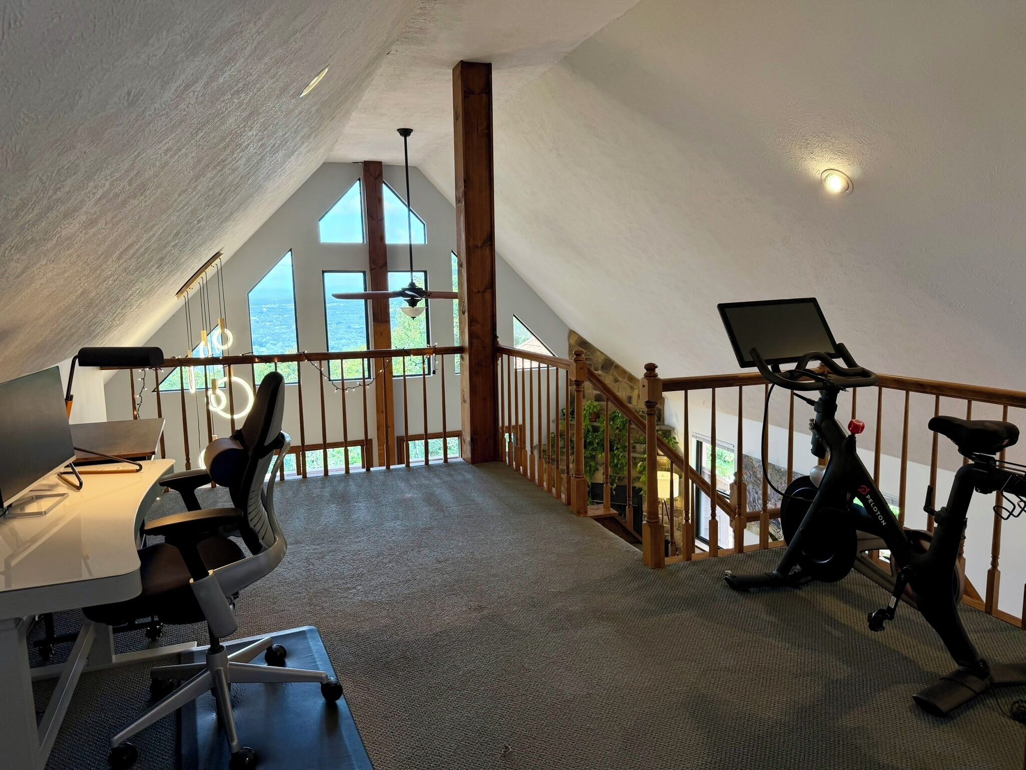 Habitación amplia con techo alto, un escritorio con computadora, una bicicleta estática y vistas al exterior a través de grandes ventanales.