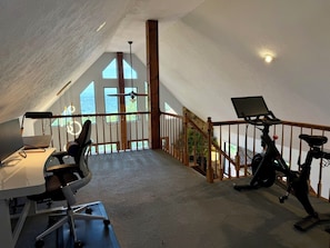 Habitación amplia con techo alto, un escritorio con computadora, una bicicleta estática y vistas al exterior a través de grandes ventanales.