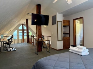 Un dormitorio con una cama grande, un escritorio con silla, un televisor sujeto a una viga de madera y vistas al exterior a través de una ventana.
