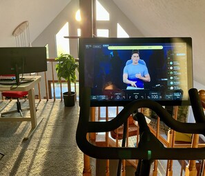 Una habitación con un monitor de computadora mostrando una interfaz de videollamada, un escritorio con teclado, una silla y una planta en maceta.