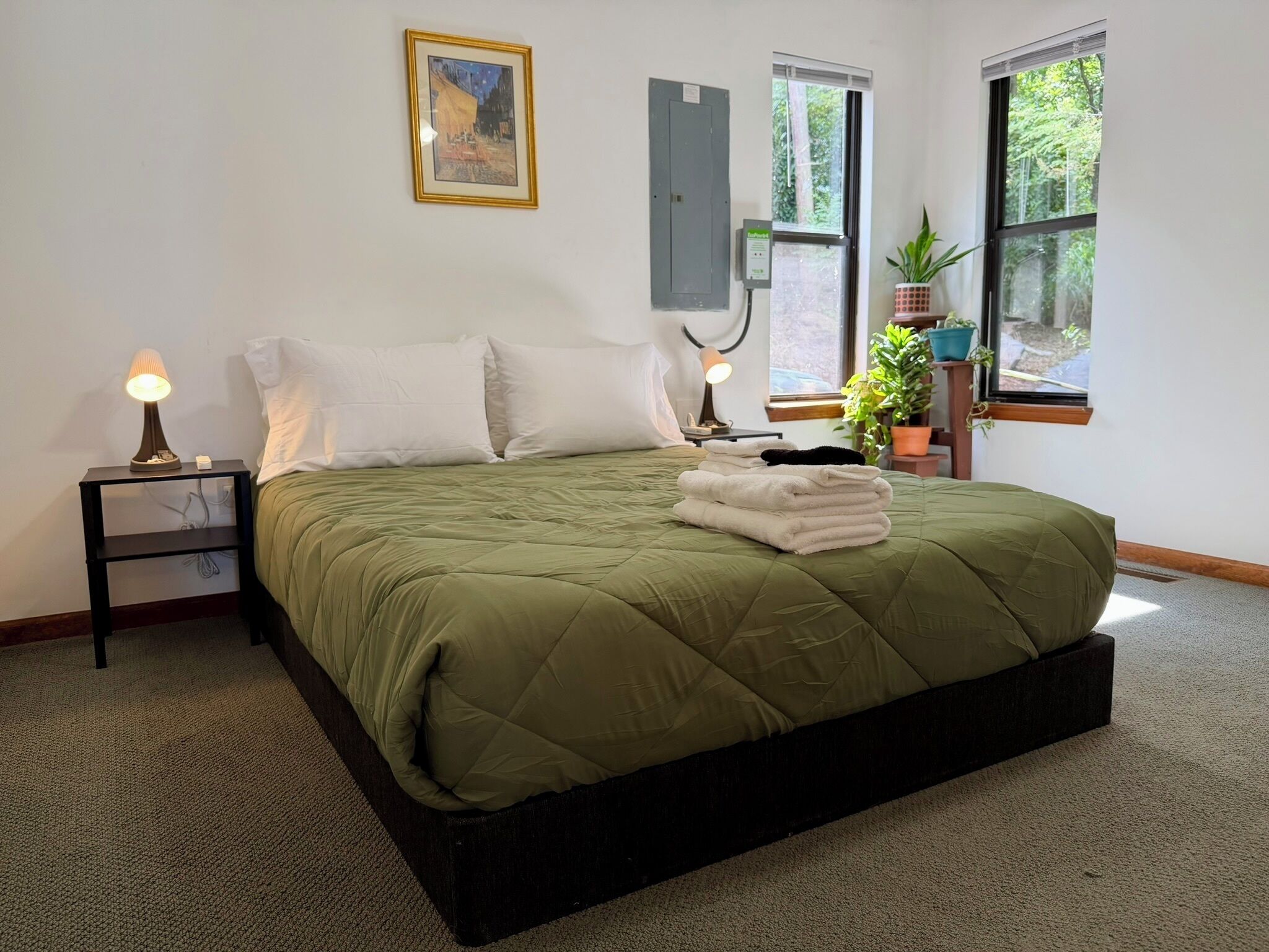 Un dormitorio con cama, dos lámparas de noche, un cuadro enmarco, una ventana con vista a árboles y una planta en maceta.