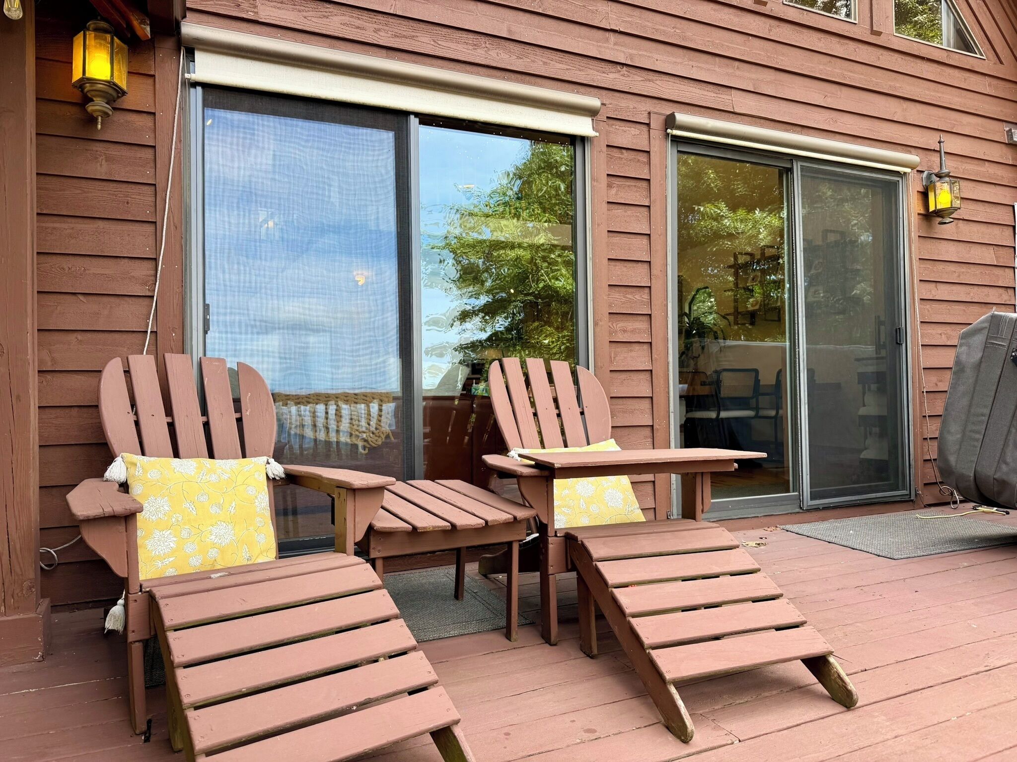 Una terraza de madera con dos sillas Adirondack y una pequeña mesa, un ventanal corredero y una ventana con vista a árboles.