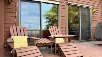 Una terraza de madera con dos sillas Adirondack y una pequeña mesa, un ventanal corredero y una ventana con vista a árboles.