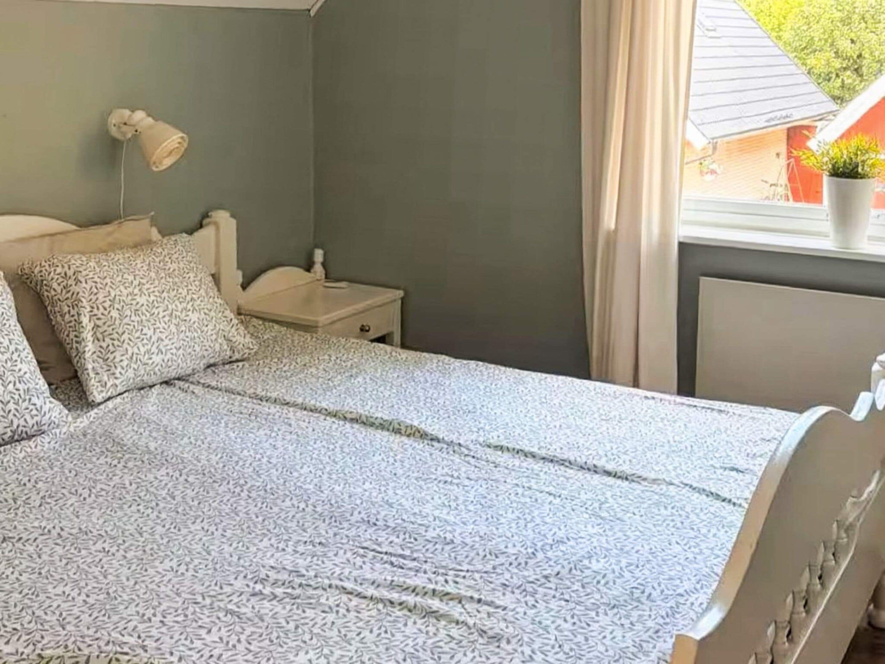 Uma cama com um cobreleito estampado, uma mesa de cabeceira com uma luminária e uma janela com cortinas.
