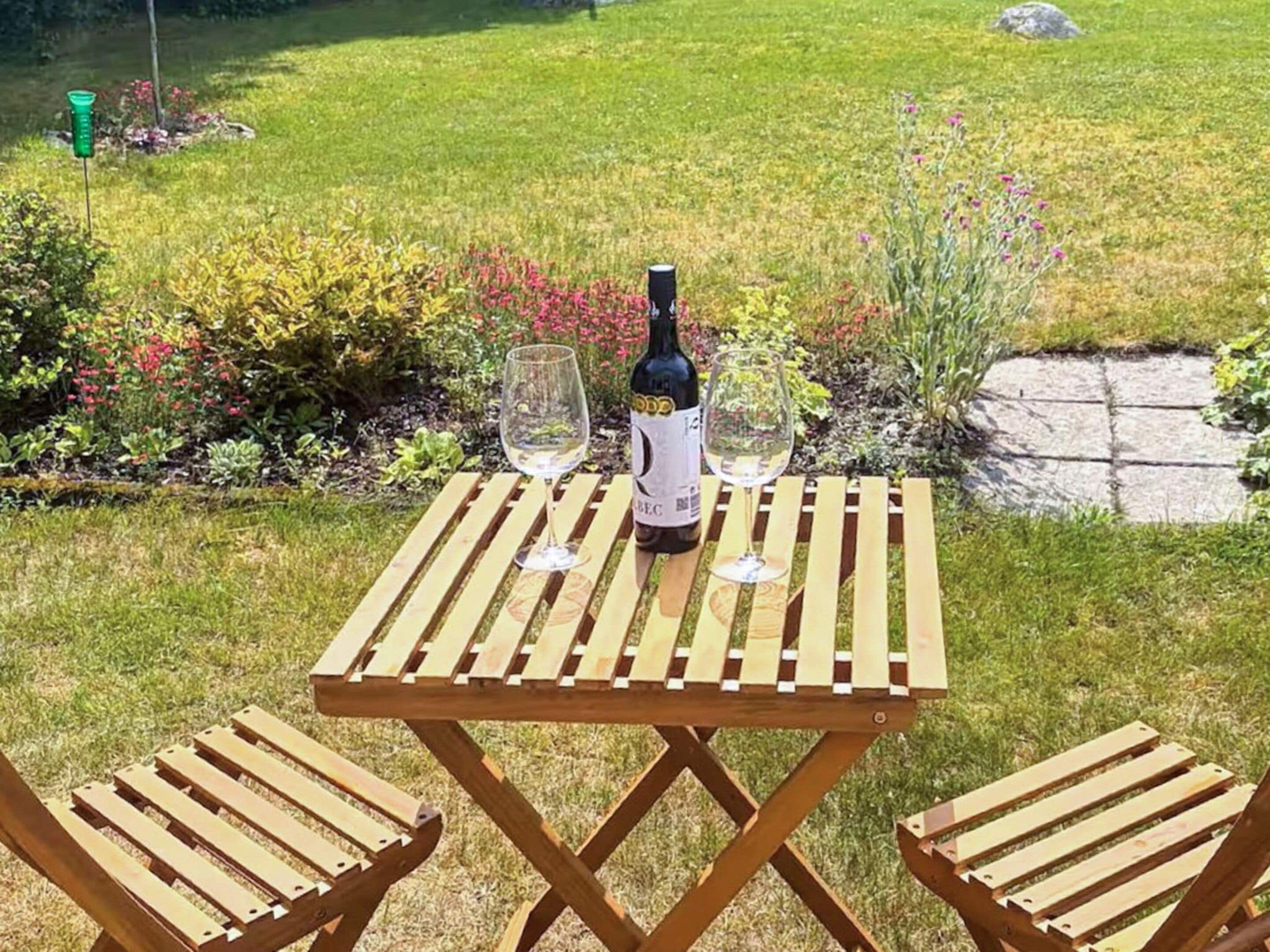 Una mesa de madera con dos sillas, una botella de vino y dos copas de vino sobre un césped con un jardín al fondo.