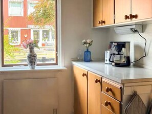 A kitchen with wooden cabinets, a window with a view of a building outside, a vase with flowers on the windowsill, and a coffee maker on the counter. - 5 Person Holiday Home in Brakne-hoby (Brakne-Hoby)
