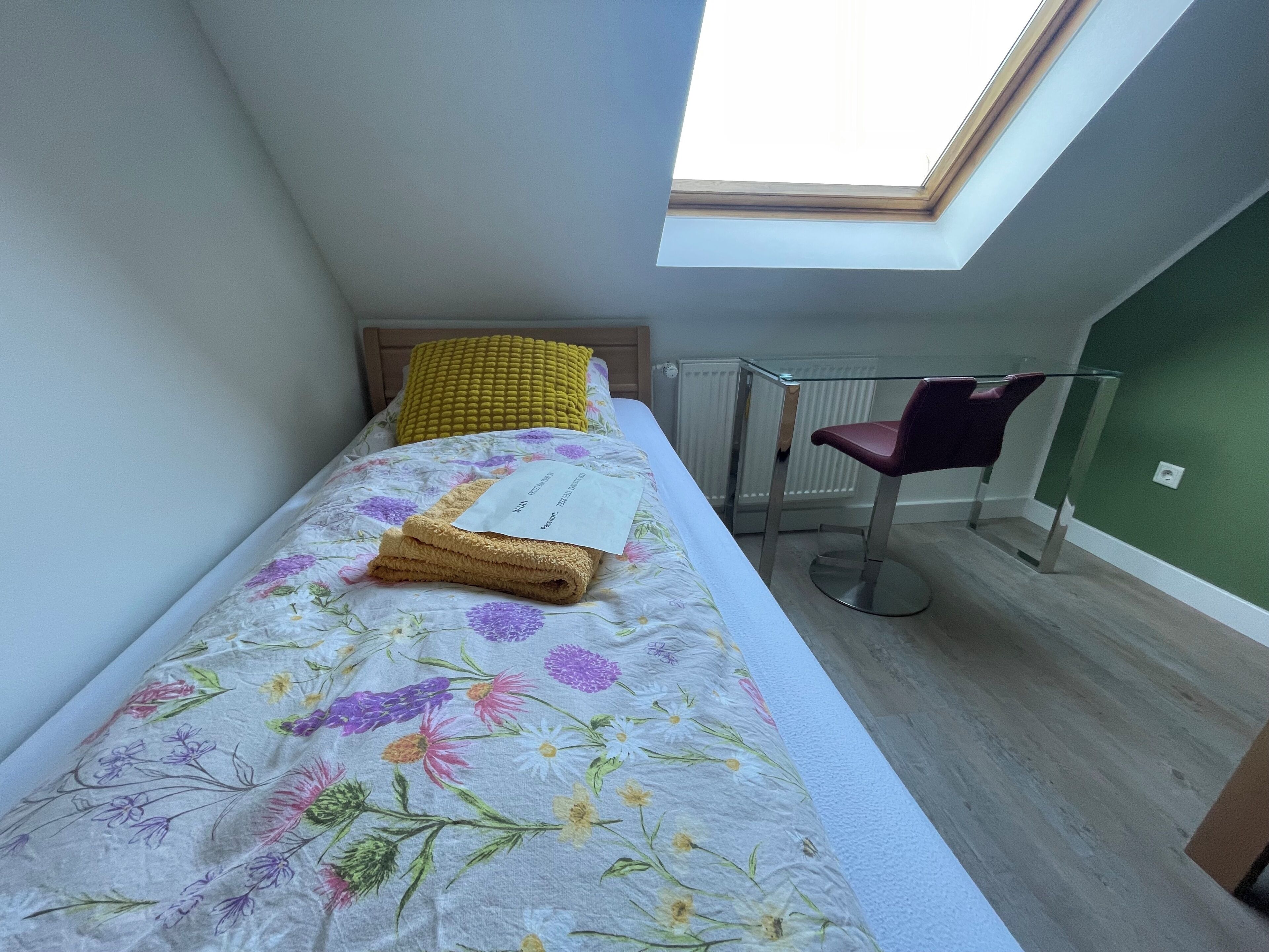 Un lit simple avec une couvre-lit à motifs floraux, un oreiller jaune et une serviette pliée. On y trouve une chaise avec un coussin foncé, un radiateur et une lucarne au-dessus du lit.