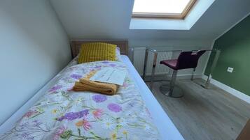 Un lit simple avec une couvre-lit à motifs floraux, un oreiller jaune et une serviette pliée. On y trouve une chaise avec un coussin foncé, un radiateur et une lucarne au-dessus du lit.