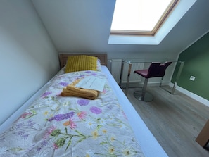Ein Einzelbett mit floralem Bettbezug, einem gelben Kissen und einem gefalteten Handtuch. Ein Stuhl mit dunkler Sitzkissen, ein Heizkörper und ein Oberlicht über dem Bett.